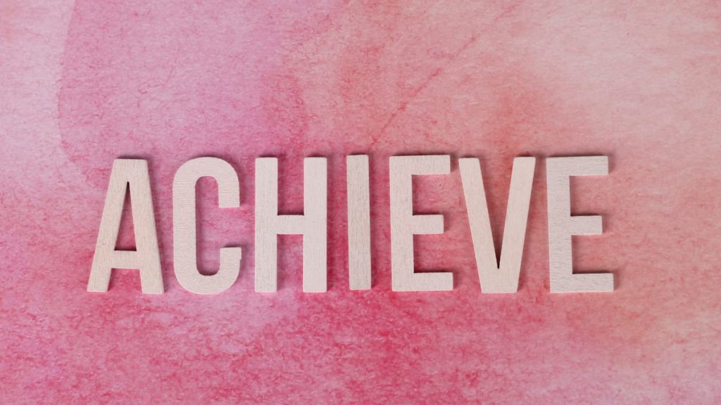 The word 'Achieve' in wooden letters on a textured pink background, symbolizing success and motivation.